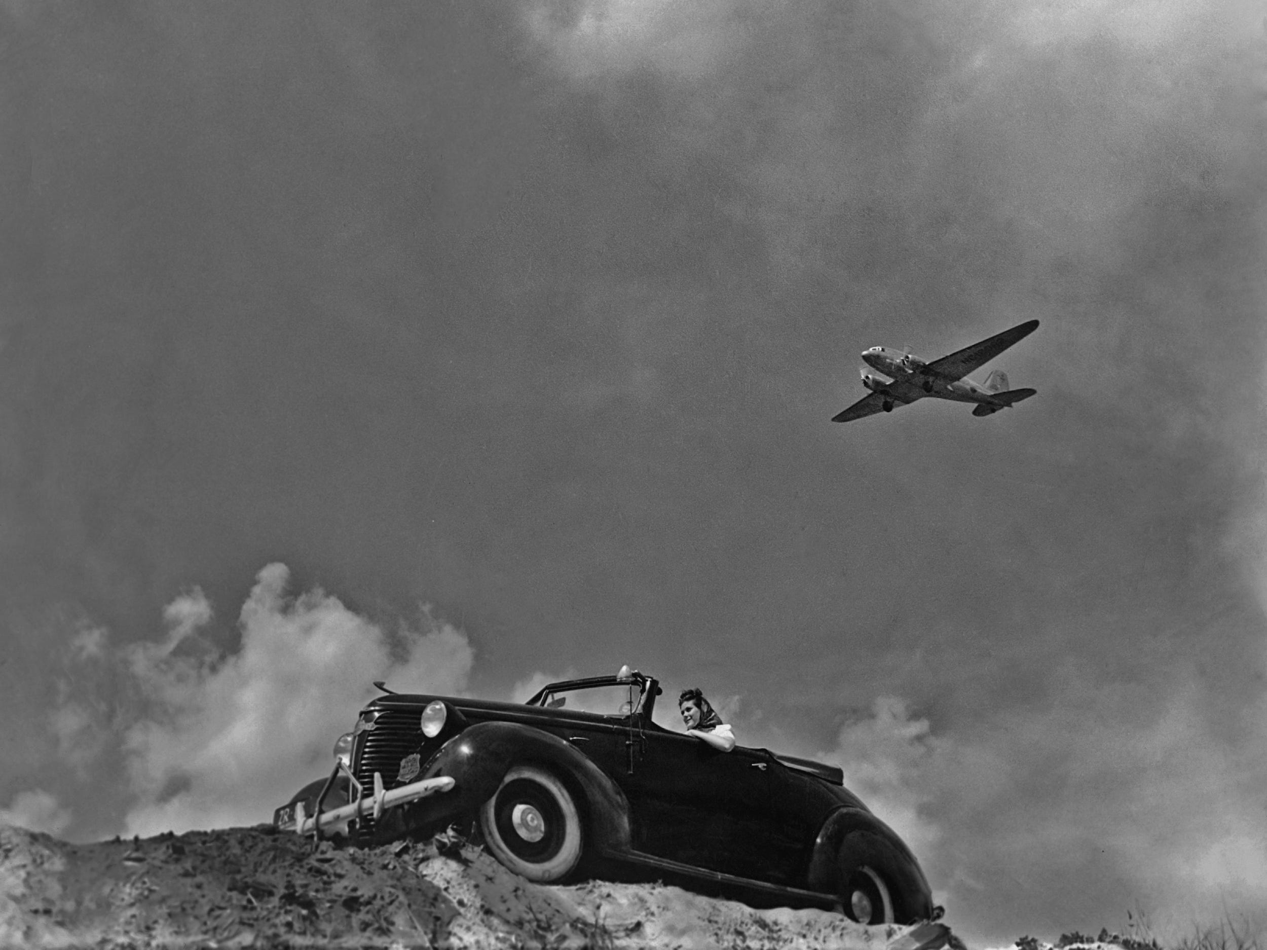 A woman sitting in a convertible car as an airplane flies overhead circa 1950