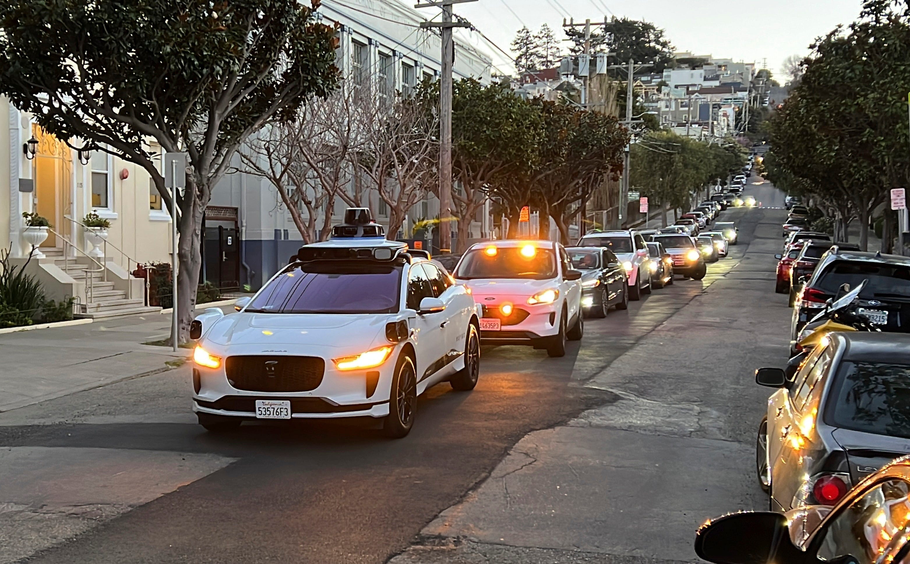 a white car blocks a line of cars waiting behind it on a city street. It's a Waymo self-driving taxi.