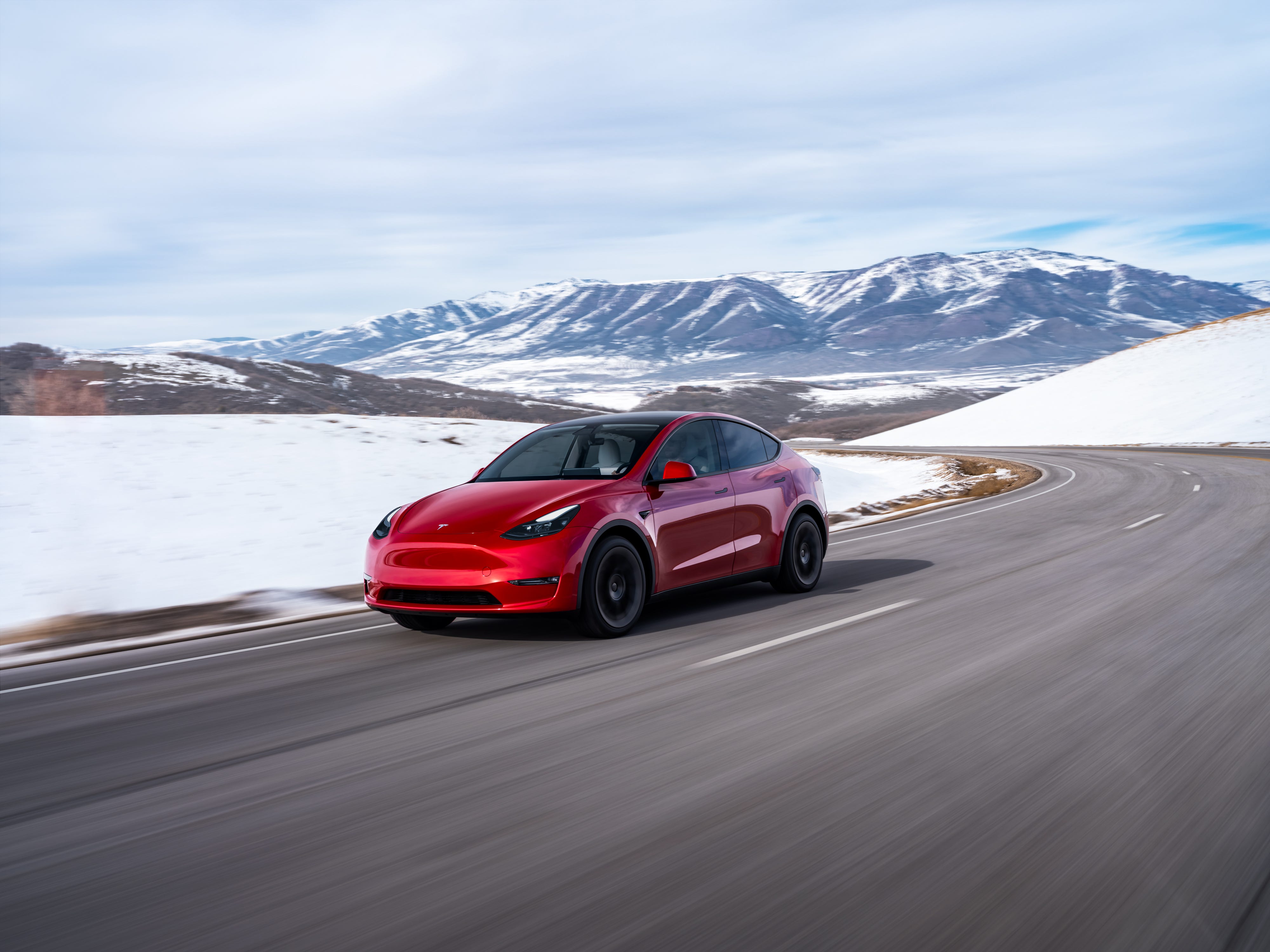 A red Tesla Model Y SUV drives down a road with snow and mountains in the background.